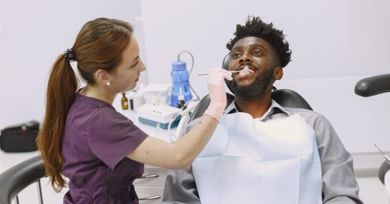 A dentist in purple scrubs and pink gloves checks a seated adult patient's teeth with a dental mirror